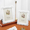 A white ornate photo frame with a rose design sits on a brown book next to a gold lamp.