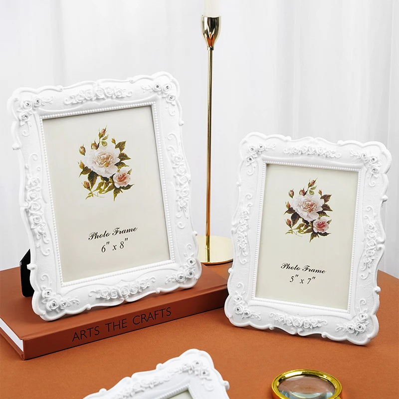 A white ornate photo frame with a rose design sits on a brown book next to a gold lamp.