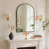 An arched wall mirror with an aluminum frame hangs above a marble mantelpiece.