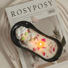 A white candle in a black silicone mold resting on a wooden surface with pink petals and a product label.