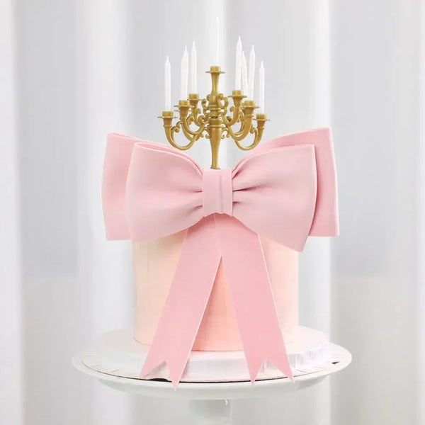 Topper de pastel con lazo de espuma rosa, candelero dorado y velas blancas sobre un pastel blanco.