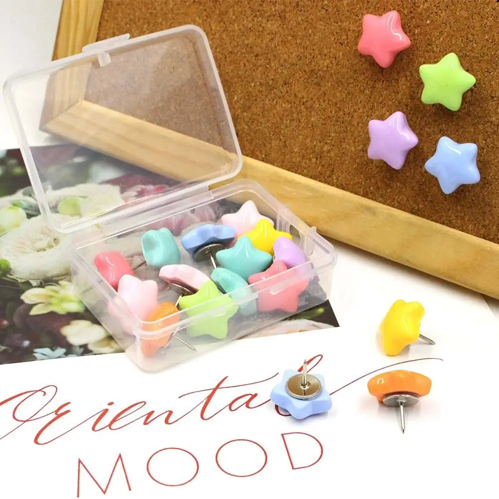 A collection of colorful pentagram thumb tacks in various shades are displayed on a cork board and in a clear plastic contain