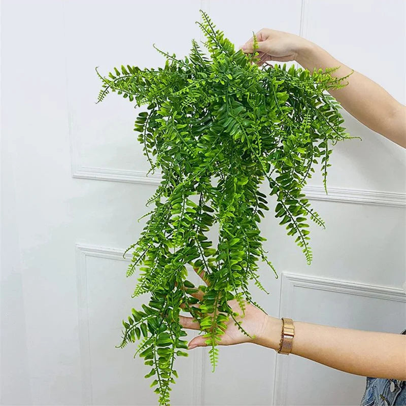 A hand is holding a lush green artificial plant vine with small leaves against a white wall.