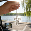 A hand holds a stainless steel praying hands cross pendant against the bright sunlight in a car window.