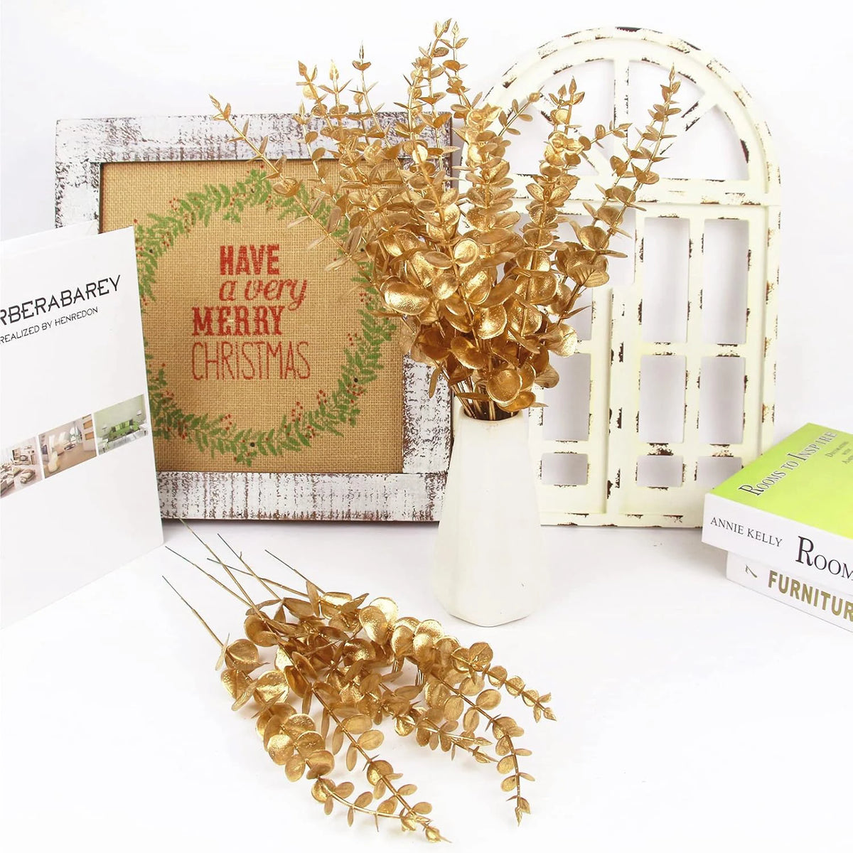 Golden artificial eucalyptus leaves arranged in a white vase against a rustic wooden frame and white door backdrop.