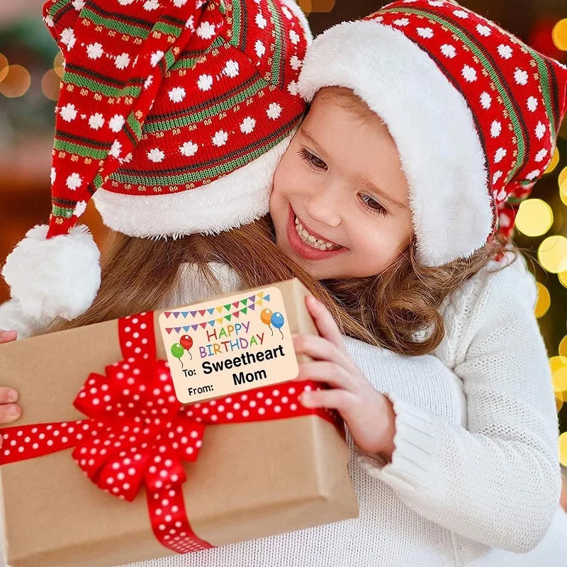 A young girl in a white sweater is smiling while holding a gift box tied with a red ribbon and a birthday tag.
