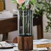 A tall glass vase with a wooden base holding green stems and a pink rose flower.