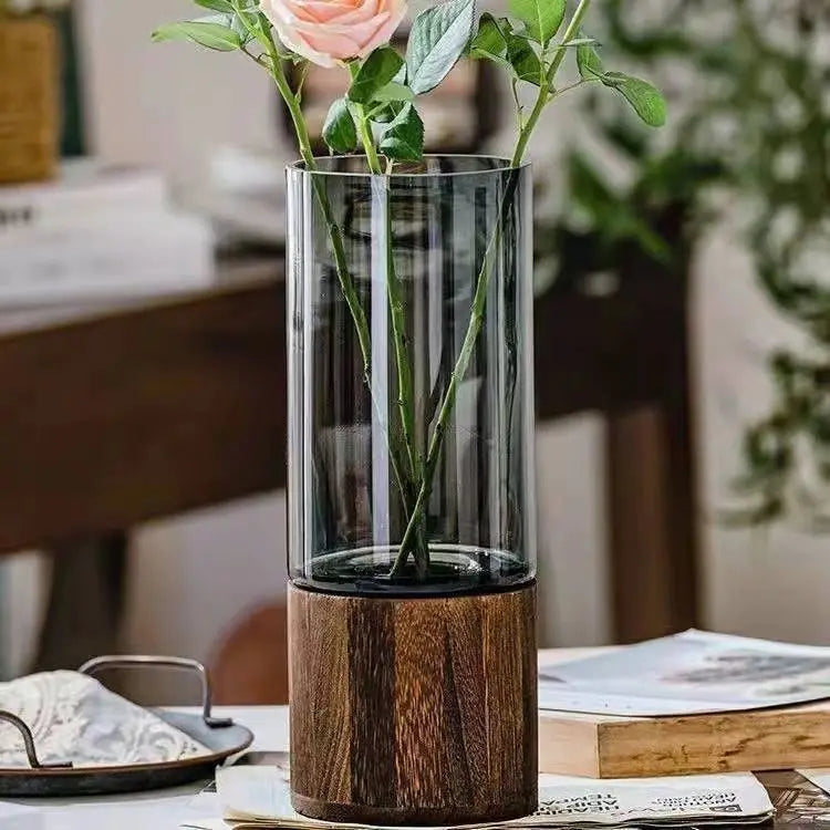 A tall glass vase with a wooden base holding green stems and a pink rose flower.