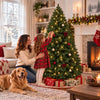 A girl and woman decorate a lit Fraser Fir artificial Christmas tree in a cozy living room setting.