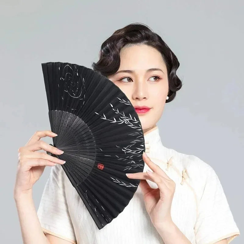 A woman holds a black vintage bamboo folding hand fan to her face against a light gray background.
