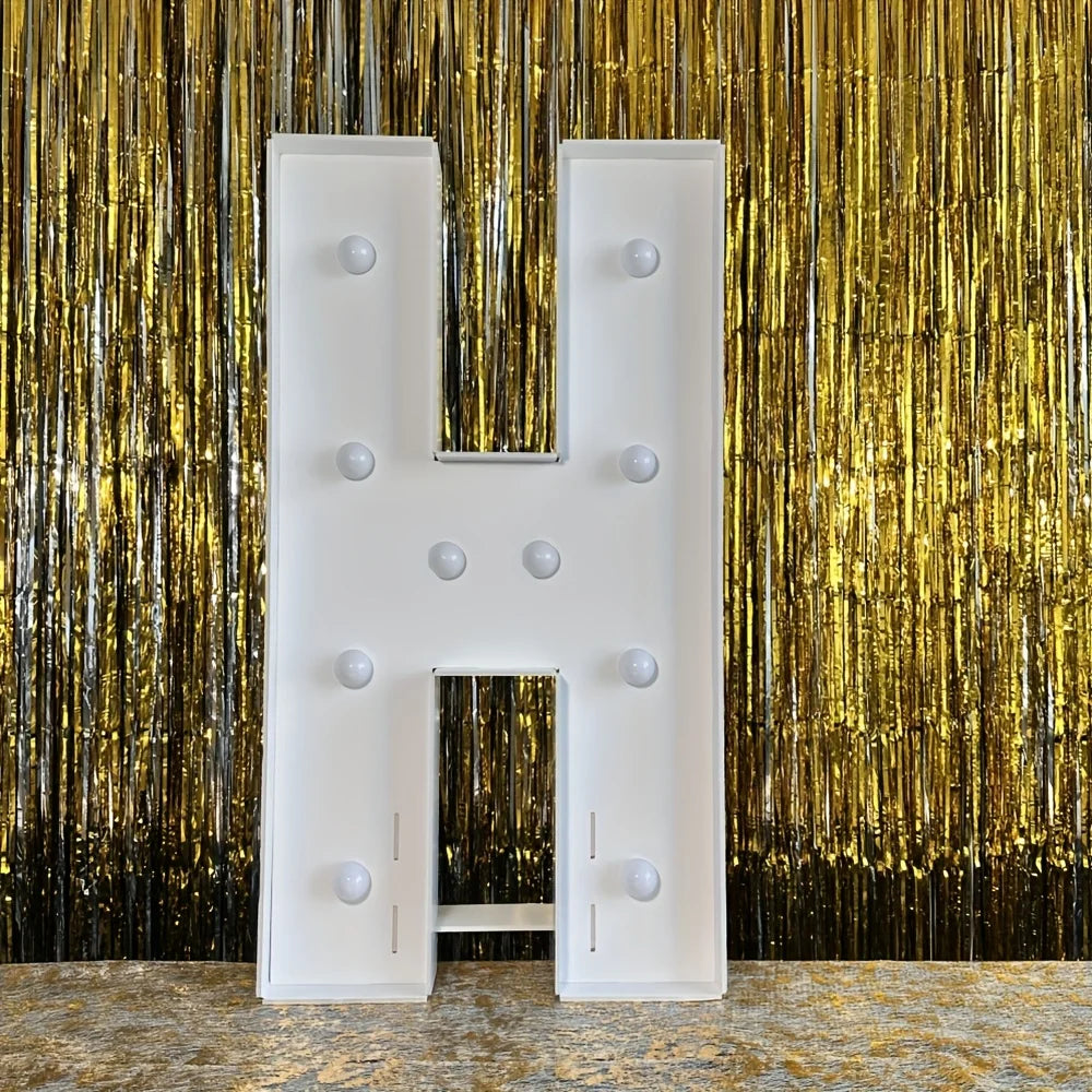 A large white marquee light-up letter H displayed against a shimmering gold curtain backdrop.