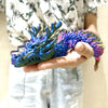 A hand holds a vibrant blue and purple articulated 3D printed dragon figurine.