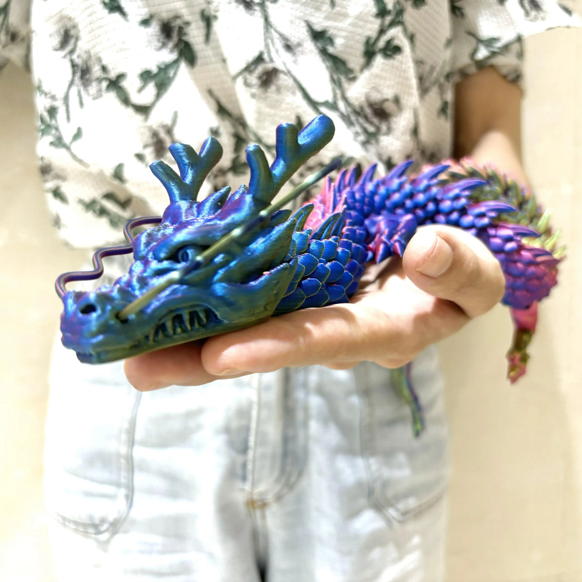 A hand holds a vibrant blue and purple articulated 3D printed dragon figurine.