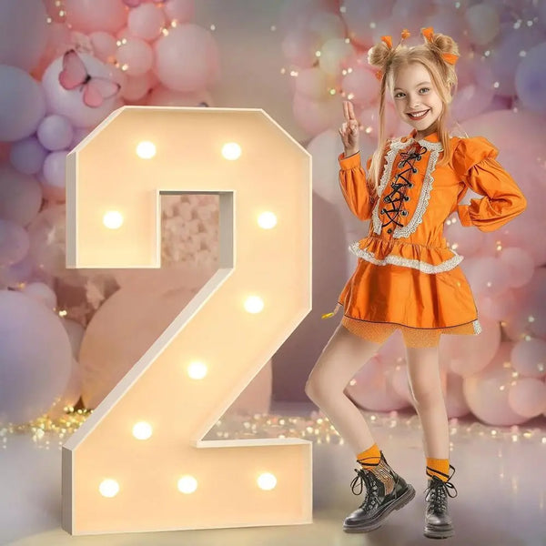 A young girl in an orange dress poses next to a large, illuminated number 2 decoration.