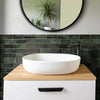 A white vessel sink sits on a wooden countertop above a dark green subway tile backsplash and a round mirror.
