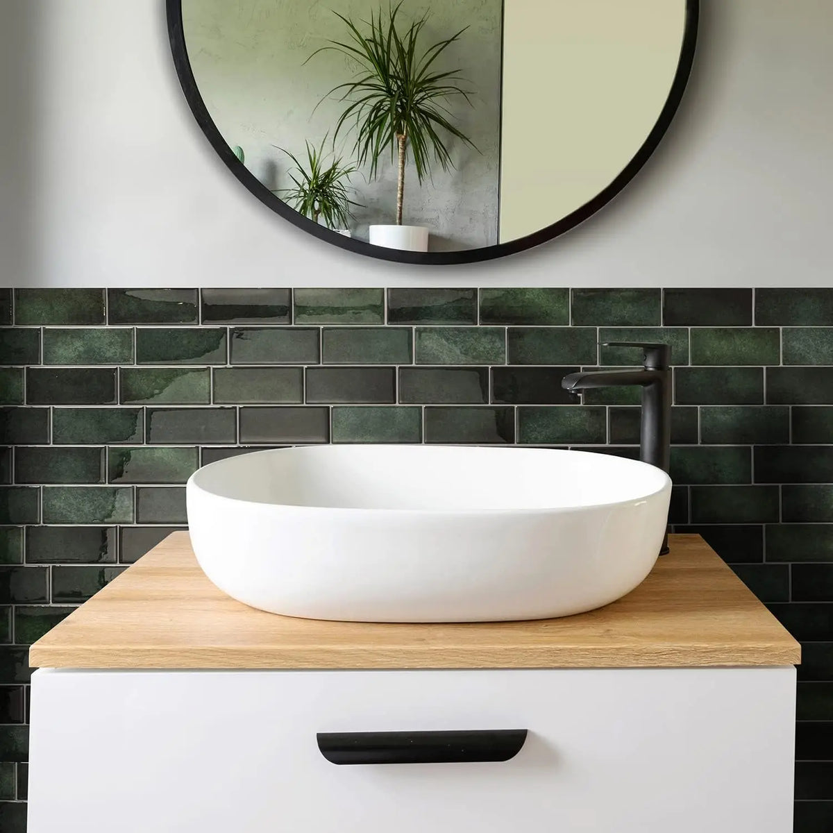 A white vessel sink sits on a wooden countertop above a dark green subway tile backsplash and a round mirror.