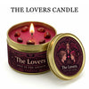 A jar of The Lovers candle with a lit wick and red wax, set against a dark pink background.
