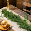 A long garland of artificial Norfolk pine greenery is displayed on a wooden table with a glass bottle and wooden utensils.
