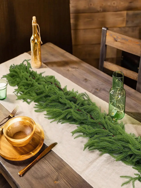 A long garland of artificial Norfolk pine greenery is displayed on a wooden table with a glass bottle and wooden utensils.