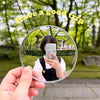 A hand holds a round convex mirror reflecting a person taking a selfie in a park setting.
