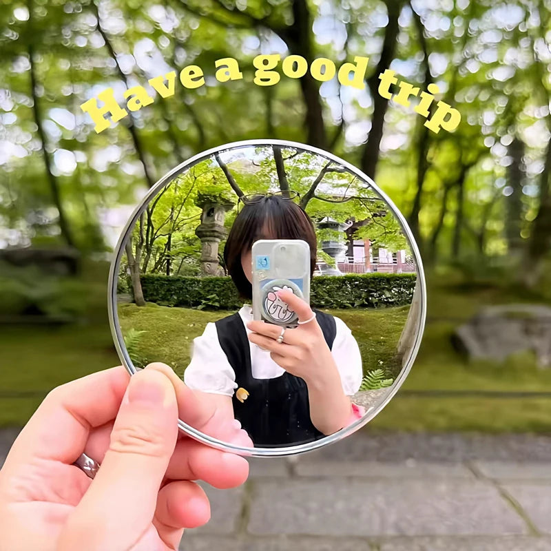 A hand holds a round convex mirror reflecting a person taking a selfie in a park setting.
