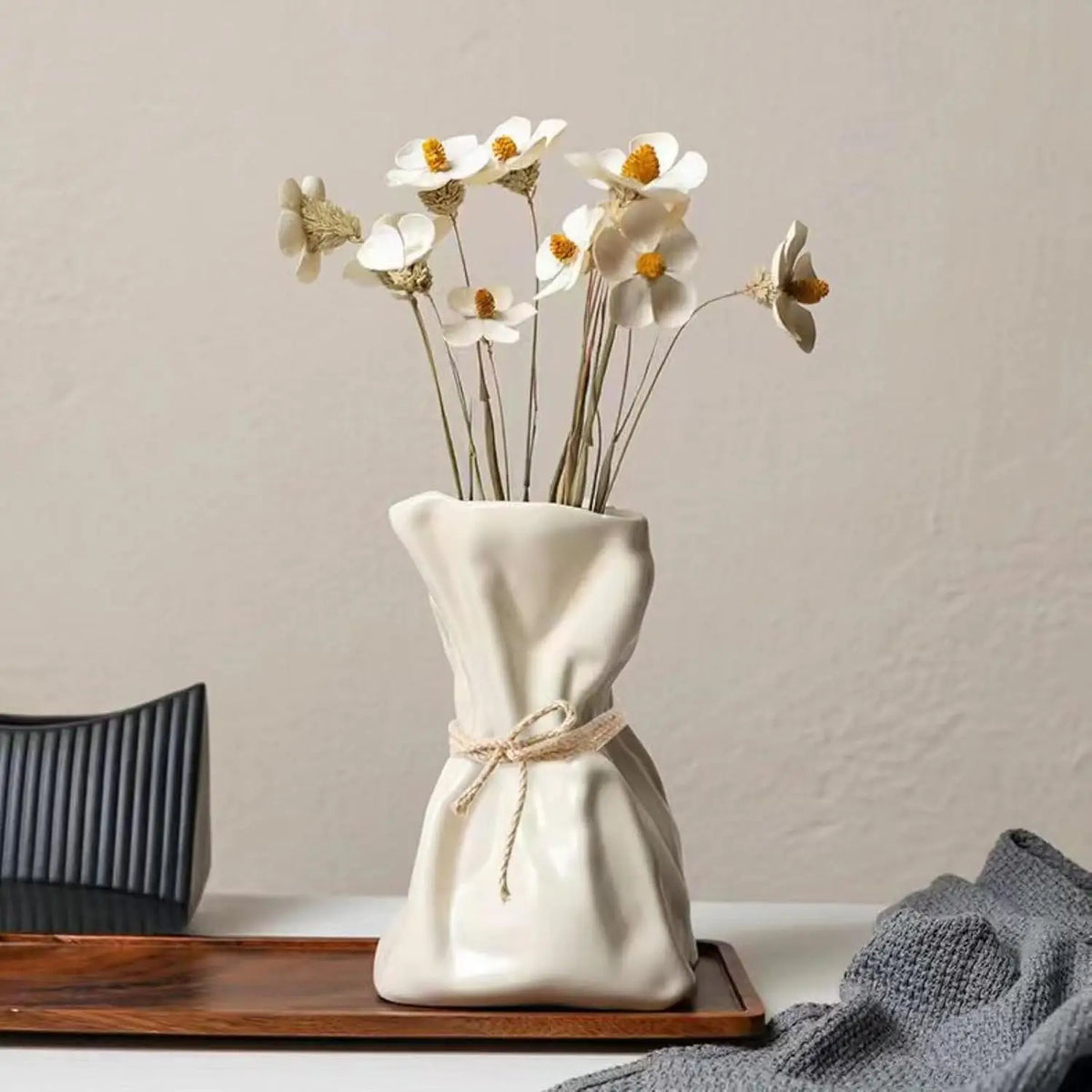 A white ceramic money bag shaped vase holds several delicate white and yellow flowers.