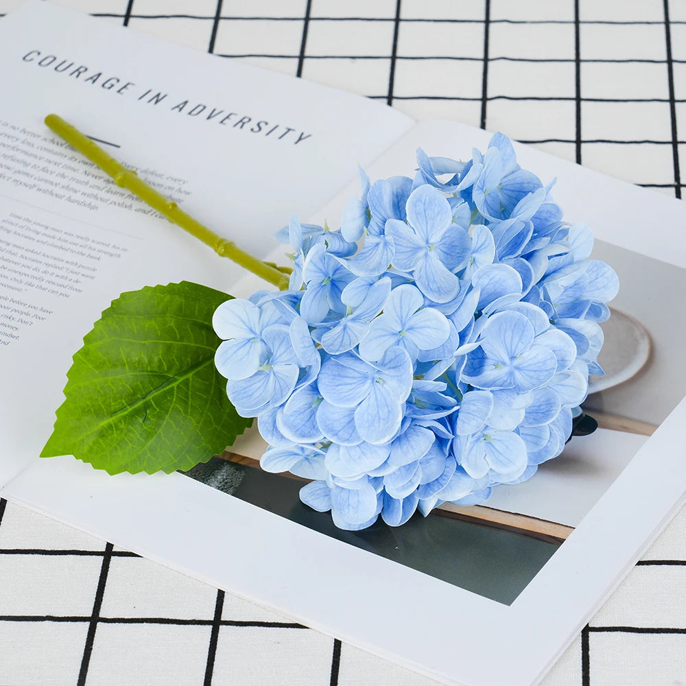 A cluster of light blue artificial hydrangea flowers rests on a white card next to green leaves.