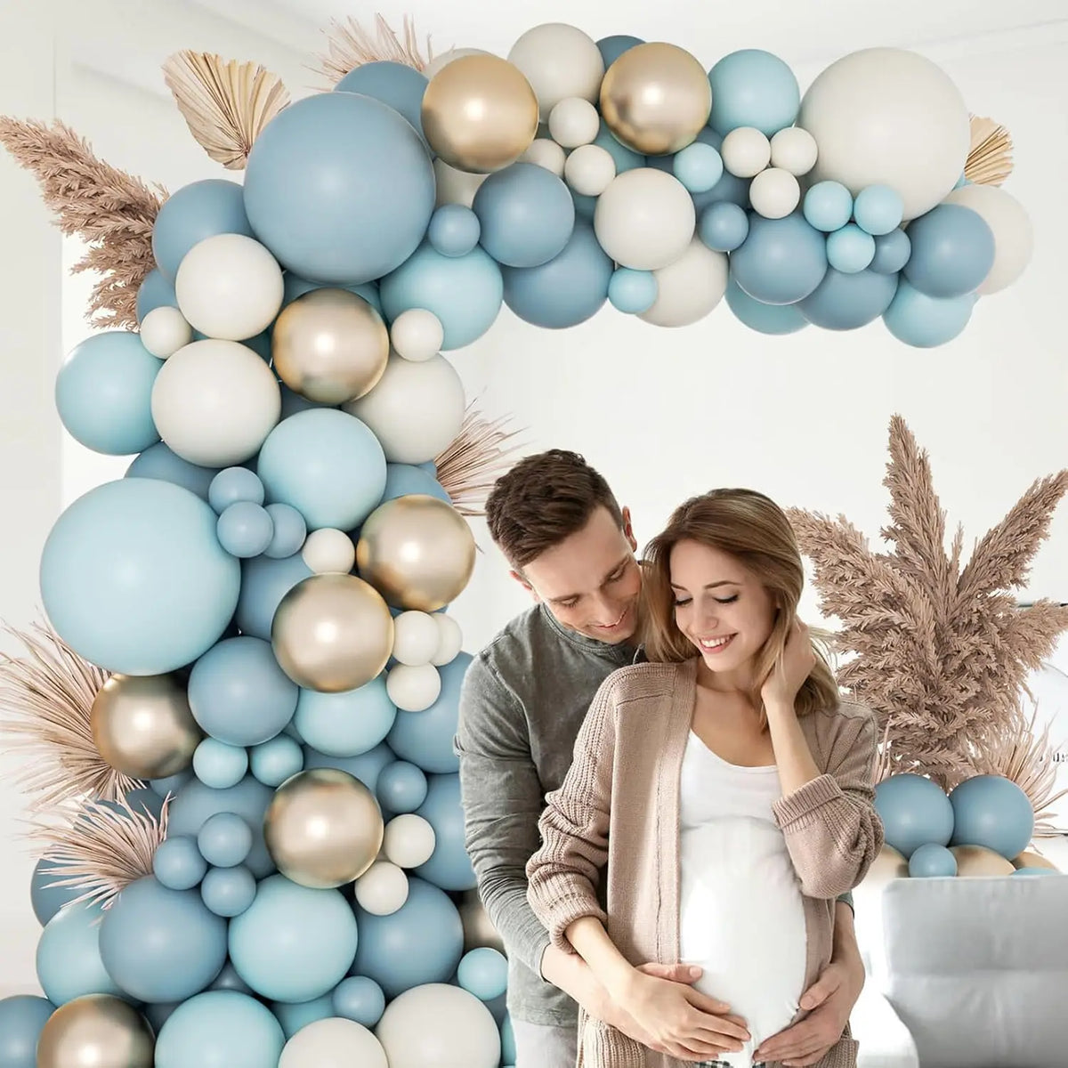 A pregnant woman is embraced by a man in front of a blue and gold balloon garland arch.