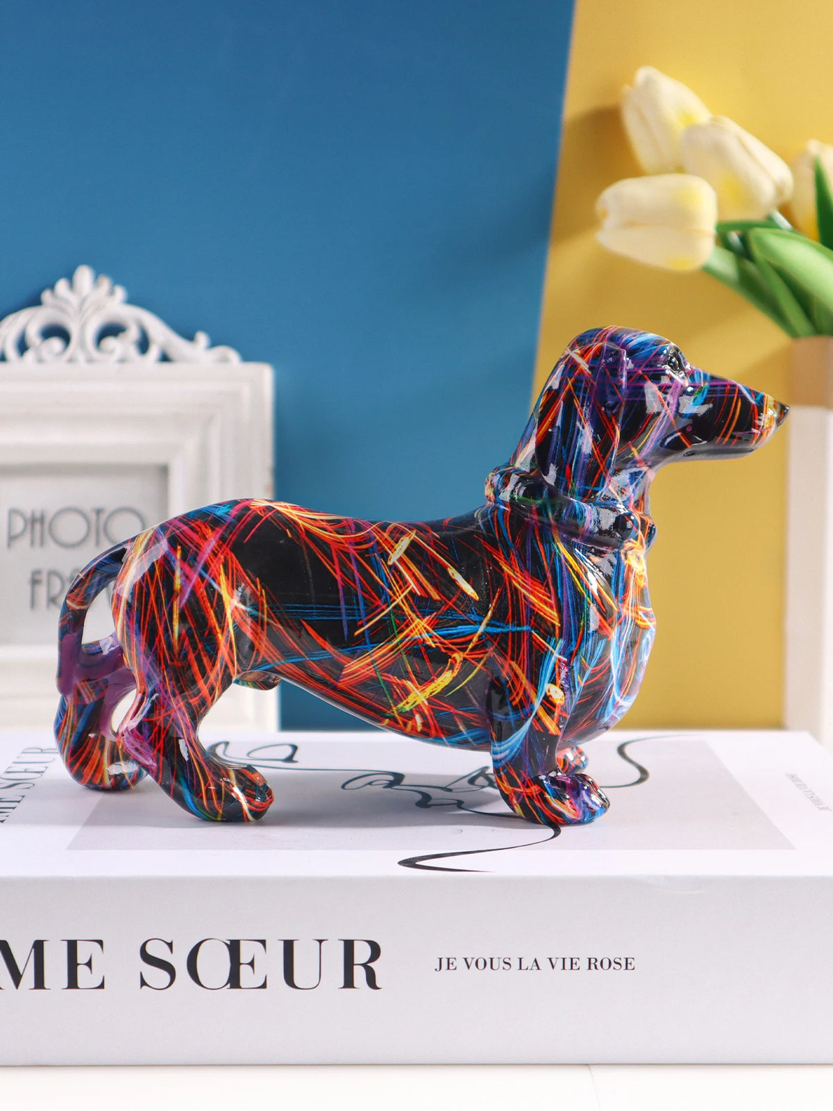 A colorful resin figurine of a dachshund dog in various vibrant colors standing on a white box.