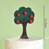 A small, dark green tree topper with red fruit decorations stands on a white cake.