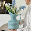 A hand holds a modern blue ceramic tabletop vase filled with white and blue flowers.