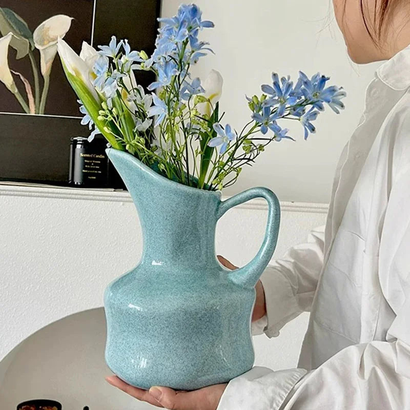 A hand holds a modern blue ceramic tabletop vase filled with white and blue flowers.