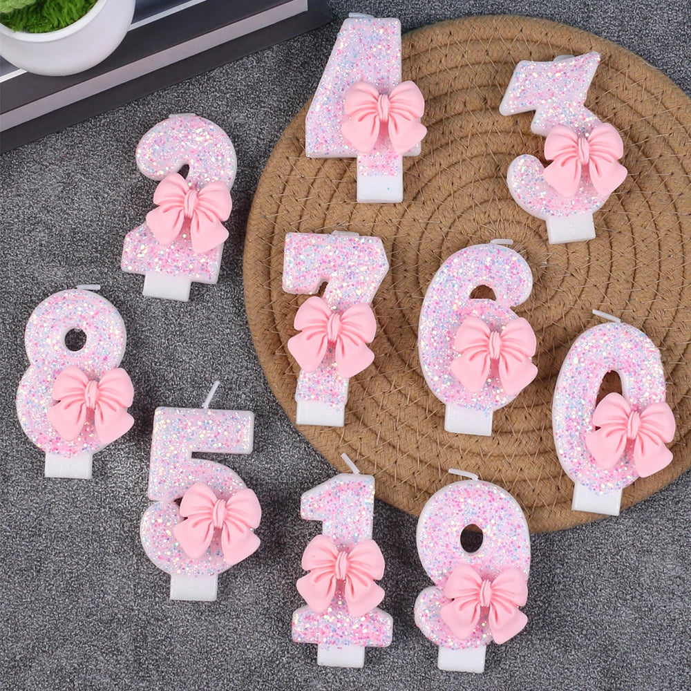 Glitter number candles shaped like 0 through 9 with pink bows arranged on a gray surface.