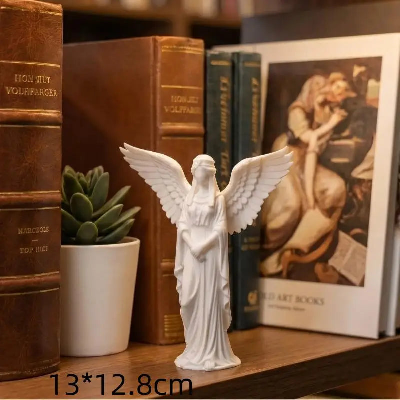 A white praying angel figurine with outstretched wings standing in front of old books and a framed artwork.