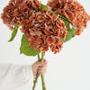 A hand holds a cluster of dried, reddish-brown hydrangea silk flower branches.