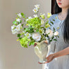 A woman holds an elegant white ceramic vase filled with fresh white and light green flowers.