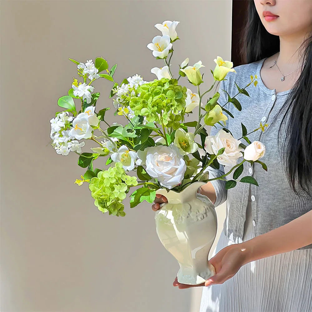A woman holds an elegant white ceramic vase filled with fresh white and light green flowers.