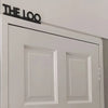 Una puerta blanca con un letrero negro que dice THE LOO sobre dos paneles rectangulares.