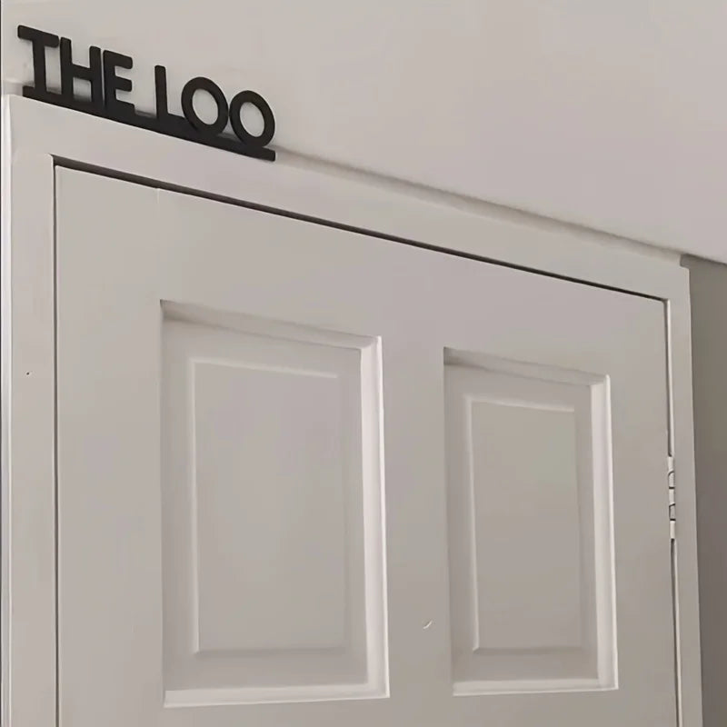 Una puerta blanca con un letrero negro que dice THE LOO sobre dos paneles rectangulares.