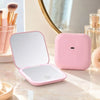 A pink portable folding LED makeup mirror with a reflective surface is displayed on a marble surface.