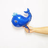 A hand is holding a blue foil balloon shaped like a dolphin with a light blue fin against a white background.