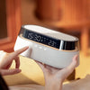 A hand holds a white wireless charging speaker lamp with a digital display and controls in a softly lit indoor setting.