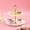 A two-tier golden serving stand holds various desserts including cake slices and small pastries on a pink background.