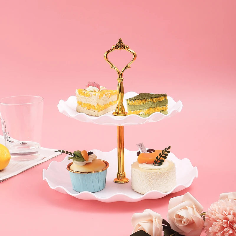 A two-tier golden serving stand holds various desserts including cake slices and small pastries on a pink background.