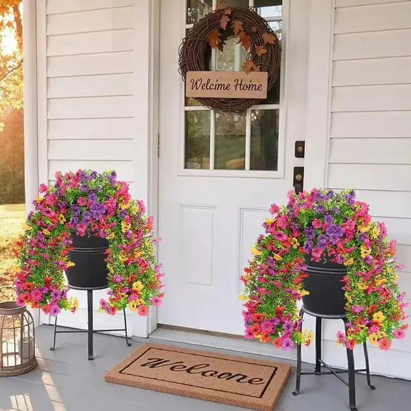 A wreath of artificial flowers in pink, purple, and yellow surrounds a black pot on a porch with a welcome mat.