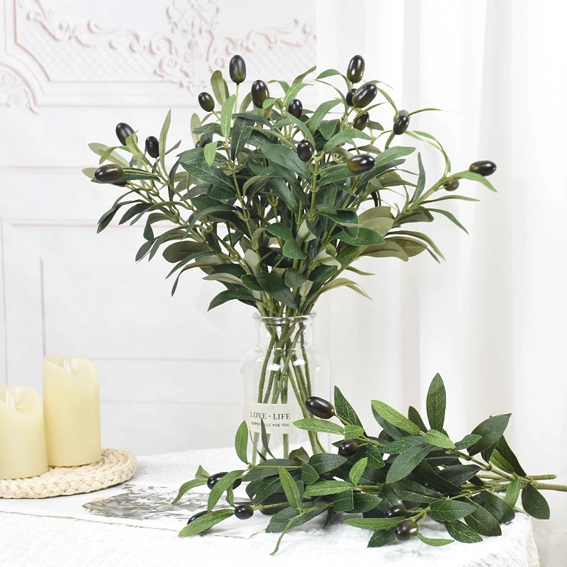 A green artificial olive leaf branch displayed in a clear glass vase on a white surface.