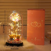 A glass dome containing a glowing golden rose, displayed next to its pink box.