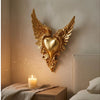 Ornate winged heart wall decor made of gold-toned metal mounted on a neutral wall.