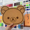 A hand holds a round brown felt cartoon bear wall decor panel with a simple face and ears.