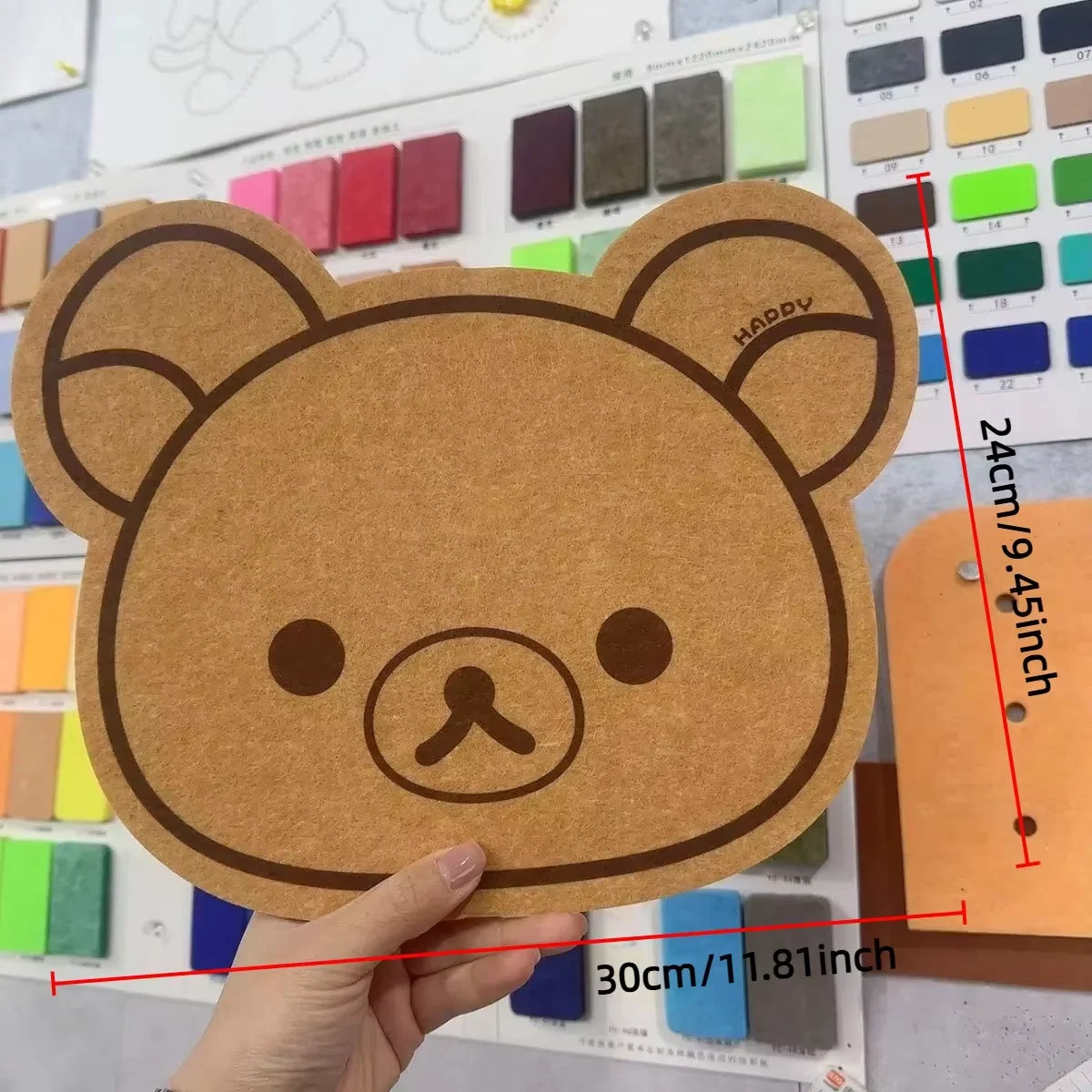 A hand holds a round brown felt cartoon bear wall decor panel with a simple face and ears.